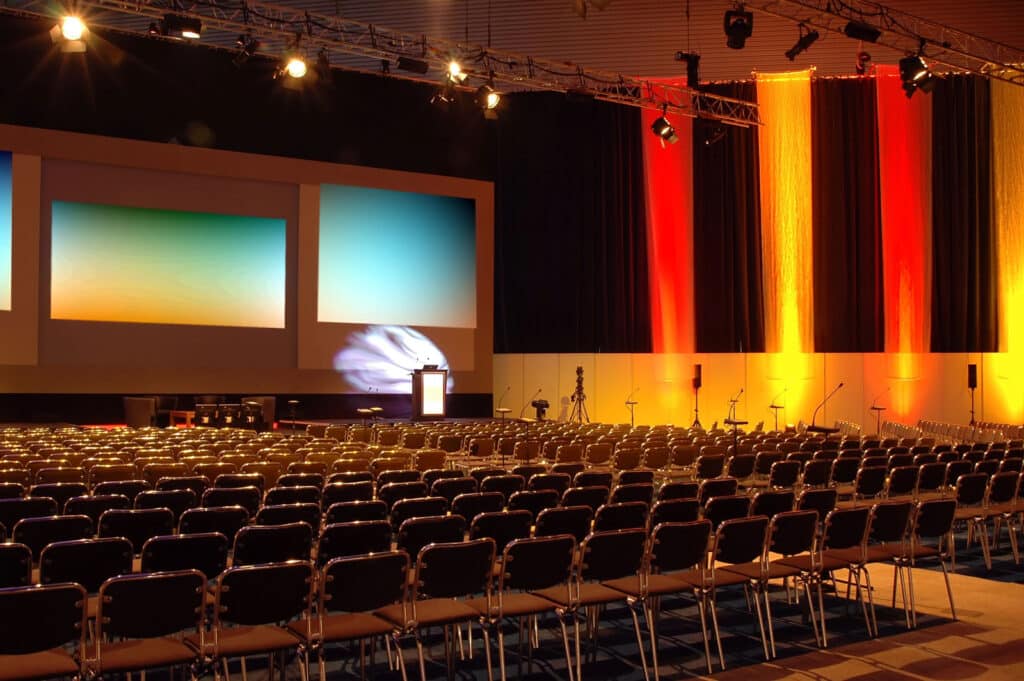 Empty auditorium with rows of chairs facing a stage with three large projection screens and colorful stage lighting.