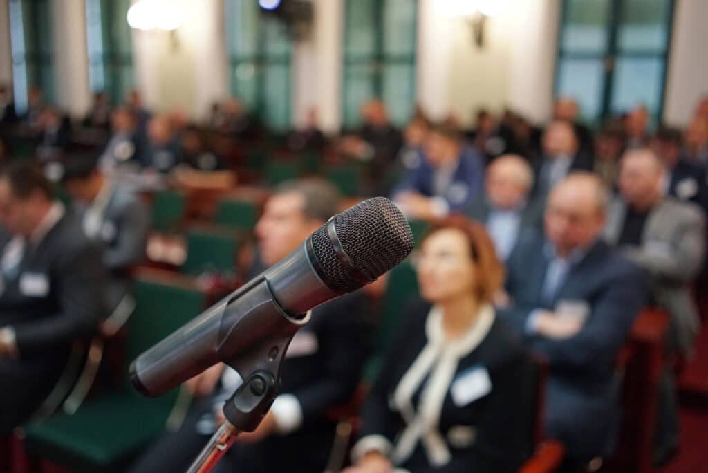 Microphone in foreground with blurred business audience seated in background.